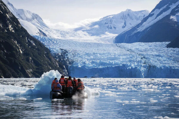 Ein Schlauchboot fährt durch eisiges Meer, im Hintergrund ein Gletscher, der zwischen zwei Bergen verläuft.