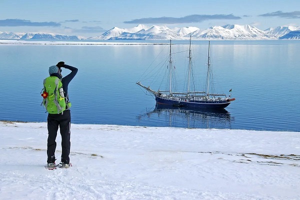 Eine Person steht im Schnee an der Küste, im Hintergrund sieht man das Meer, ein Schiff und Eisberge