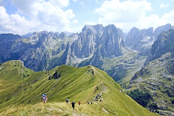 Ausblick auf Gebirge im Hintergrund und grünem Berg im Vordergrund, auf dem ein Wanderweg mit Menschen zu sehen ist